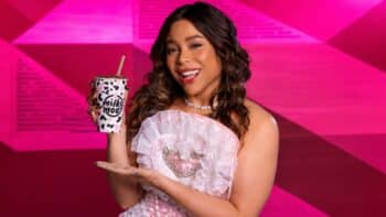 Milky Moo com Blogueirinha e novo sabor Amarga Mulher sorrindo em cenário rosa segurando um copo da Milky Moo com estampa de vaquinhas, usando vestido claro e colar de pérolas.