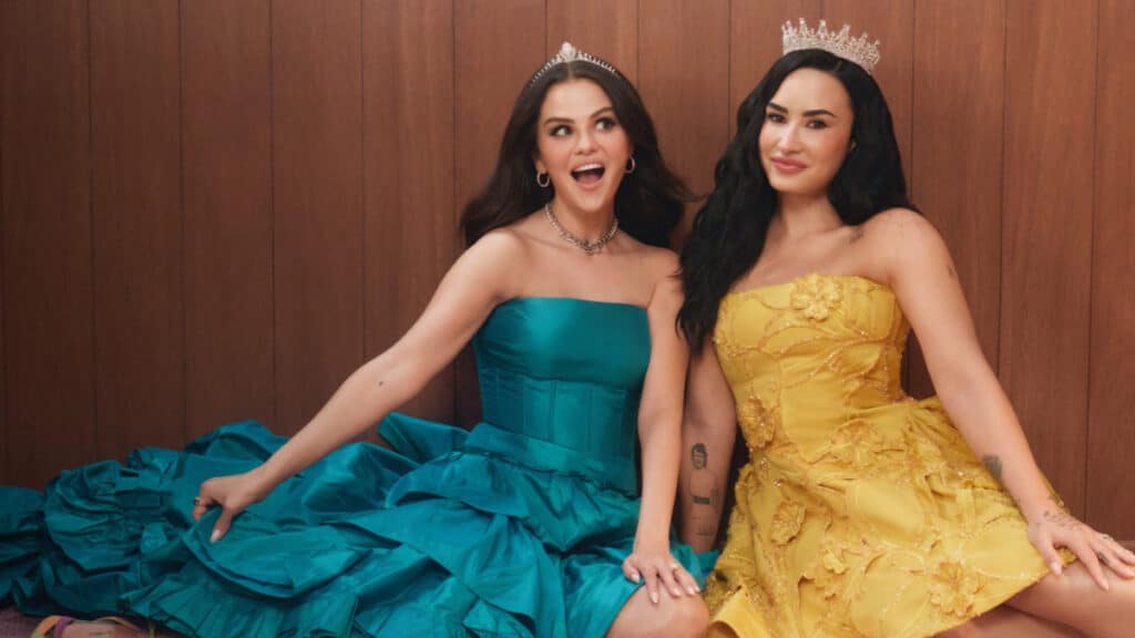 Selena Gomez e Demi Lovato na campanha Rare Beauty Selena Gomez e Demi Lovato posam juntas em campanha da Rare Beauty, usando vestidos azul e amarelo, em cenário com fundo de madeira.