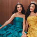 Selena Gomez e Demi Lovato posam juntas em campanha da Rare Beauty, usando vestidos azul e amarelo, em cenário com fundo de madeira.
