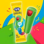 Picolé Fini Minhocas em embalagem e palito, com cobertura verde e imagem do personagem azul, sobre fundo colorido