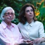 Fernanda Montenegro e Fernanda Torres sentadas juntas em campanha do TikTok, em cenário externo com flores